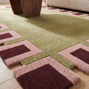 Pu&ograve; includere: Un tappeto rettangolare caratterizzato da un campo centrale verde oliva testurizzato, bordato da un motivo geometrico di quadrati alternati rosa chiaro e bordeaux scuro. Il tappeto presenta una struttura a pelo spesso e denso.
