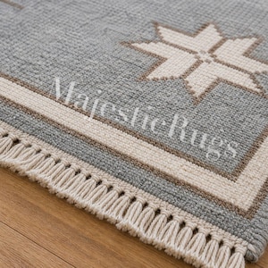 May include: Close-up of a gray rug with a star design. The rug has a cream-colored border and fringe. The star is cream-colored. The rug is on a wooden surface. The text "Majestic Rugs" is visible on the rug.