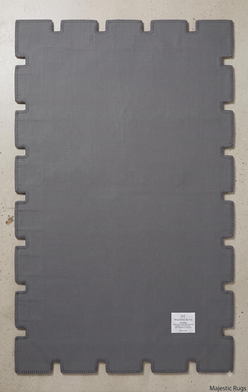Pu&ograve; includere: Un tappeto rettangolare grigio scuro caratterizzato da un bordo dentellato a forma di pezzo di puzzle con cuciture a festone. Un'etichetta rettangolare bianca nell'angolo in basso a destra riporta il testo MAJESTIC RUGS, Hand Tufted, Cotton Canvas Backing, Made in India.