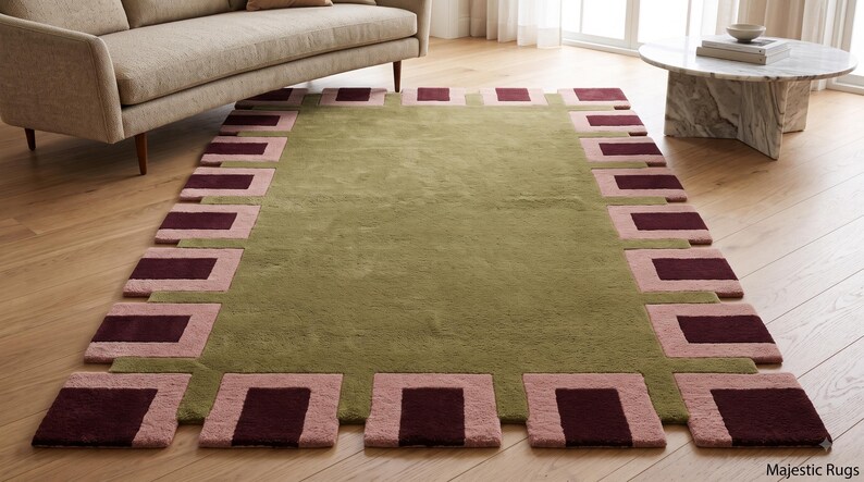 Pu&ograve; includere: Un tappeto rettangolare con un campo centrale verde oliva a tinta unita, circondato da un bordo geometrico di quadrati e rettangoli che alternano il rosa e il bordeaux scuro. Il tappeto presenta una superficie a pelo testurizzato su un pavimento in legno.