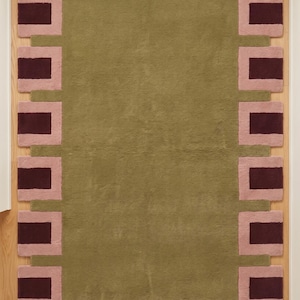 Pu&ograve; includere: Un tappeto rettangolare con un campo centrale verde oliva a tinta unita, circondato da un bordo di blocchi quadrati alternati in rosa e bordeaux scuro. Il tappeto presenta una superficie a pelo testurizzato su un pavimento in legno chiaro.