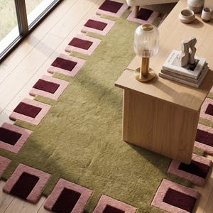 Pu&ograve; includere: Un tappeto rettangolare con un pannello centrale in verde oliva tinta unita, circondato da un bordo di blocchi quadrati che si alternano tra il rosa e il bordeaux scuro. Il tappeto presenta una superficie a pelo testurizzato su pavimento in legno chiaro.
