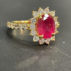 May include: A gold ring with a large, oval ruby centre stone, encircled by a halo of smaller, round diamonds. The band is also set with diamonds. The ring is displayed against a dark backdrop.
