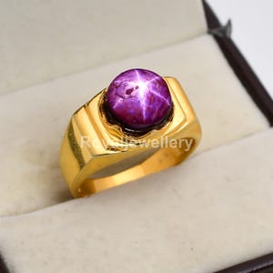 Könnte beinhalten: Ein goldfarbener Ring mit einem großen, runden, purpurroten Edelstein mit sternförmigem Muster. Der Ring hat eine rechteckige Fassung und ein dickes Band. Der Text "Rowe Jewellery" ist auf dem Ring zu sehen.