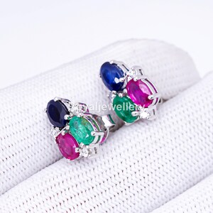 May include: A pair of silver earrings, each featuring a cluster of gemstones. The earrings showcase oval-cut sapphires, emeralds, and rubies, with small accent stones. The gemstones are set in a silver metal.