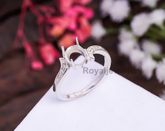 Könnte beinhalten: Ein silberner Ringrohling mit einer zentralen runden Fassung und zwei Zinken. Das Ringband ist gebogen und mit kleinen, eingebetteten, klaren Steinen versehen. Der Text "Royal Jewellery" ist sichtbar.