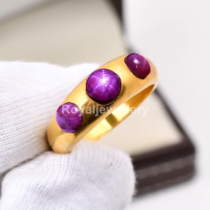 May include: A gold ring featuring three round, deep purple gemstones. The ring has a smooth, polished band and the gemstones are evenly spaced across the top. The ring is displayed in a jewelry box.