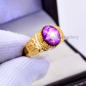 May include: A gold ring featuring a large, oval-cut, star-shaped purple gemstone. The ring has an ornate, engraved design on the band and around the gemstone setting. The ring is held by a gloved hand.