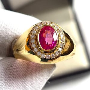 Peut inclure: Une bague en or ornée d'un grand rubis ovale entouré d'un halo de petits diamants clairs. La bague a un anneau en or poli et un design classique et élégant. Le rubis est d'une couleur rose vif.