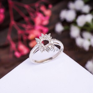 Peut inclure: Une bague en argent avec un motif floral. La bague présente un sertissage central entouré de petites pierres claires, créant une forme de fleur. Le bracelet est lisse et simple, et la bague est posée sur un fond flou.