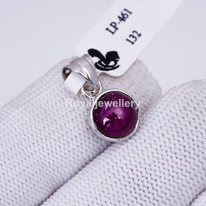 Pode incluir: Um pingente de prata com uma pedra preciosa redonda, roxa escura. A pedra preciosa é definida em uma moldura de prata e tem um efeito de estrela. Uma etiqueta branca com o texto "LP-461 132" está anexada.