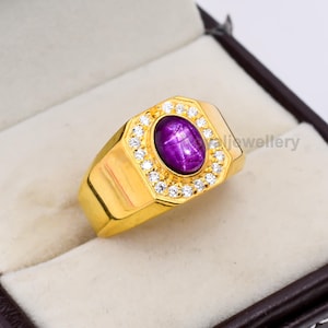 May include: A gold ring featuring a large, oval, deep purple gemstone, encircled by a halo of small, clear gemstones. The ring has a rectangular face and a thick, polished band. The ring is presented in a brown jewellery box.