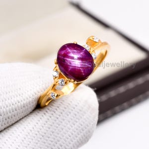 May include: A gold ring with a large, oval, deep red gemstone exhibiting a star effect. The ring is embellished with small, clear accent stones and leaf-shaped details on the band. The ring is held by a gloved hand.
