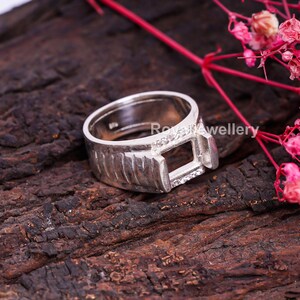 May include: A silver ring with a rectangular setting, likely for a gemstone. The ring features a textured band and small accent stones near the setting. Set against a dark, textured surface with pink floral accents.
