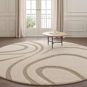 May include: A large, cream-colored, oval area rug with a repeating, swirling pattern in a lighter beige. The rug is centered in a room with a small, round wooden table and a black metal base. The room has hardwood floors and white walls.