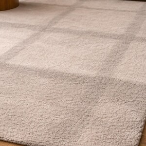 K&ouml;nnte beinhalten: Rechteckiger Teppich in hellem Beige mit dezentem Gittermuster. Der Teppich hat eine weiche, strukturierte Oberfl&auml;che und liegt auf einem Holzboden. Die neutrale Farbe erzeugt eine warme und einladende Atmosph&auml;re.