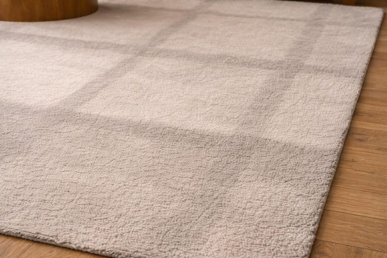 K&ouml;nnte beinhalten: Rechteckiger Teppich in hellem Beige mit dezentem Gittermuster. Der Teppich hat eine weiche, strukturierte Oberfl&auml;che und liegt auf einem Holzboden. Die neutrale Farbe erzeugt eine warme und einladende Atmosph&auml;re.