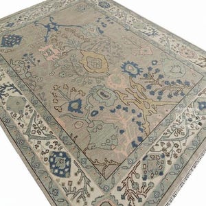 May include: A rectangular area rug with a muted color palette. The rug features a central floral design in shades of blue, pink, and yellow on a neutral background. A detailed border frames the design, adding to its decorative appeal.