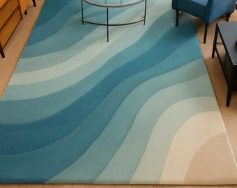 Puede incluir: Una alfombra grande con un diseño de ondas en tonos azules y beige. La alfombra está colocada en un salón, complementando el sofá y el sillón azules. El diseño de la alfombra añade un toque costero a la habitación.