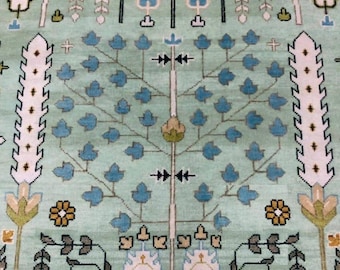 May include: A detailed, close-up view of a light green rug with an intricate tree of life design. The tree features blue leaves and a white trunk. The rug's border has floral and geometric patterns in shades of blue, white, and gold.