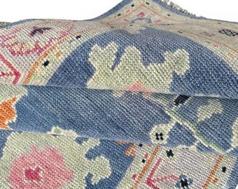 May include: Close-up of a folded area rug with a detailed pattern. The rug features a blue border with a repeating floral design in pink, orange, and green. The rug has a textured surface and a fringed edge.