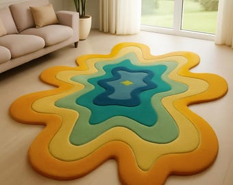 May include: A colorful, abstract rug with a unique, organic shape. The rug features concentric layers of color, transitioning from a vibrant orange outer edge to shades of yellow, green, and blue in the center. The rug is placed on a light-colored wooden floor.