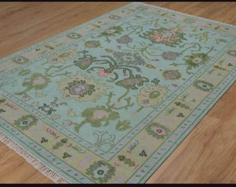 May include: Rectangular area rug with a light blue background and a floral pattern in shades of green, pink, and beige. The rug has a detailed border and fringe on one end. The rug is placed on a wooden floor.