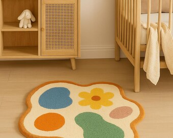 May include: A cream-colored, abstract-shaped rug with a yellow-orange border. The rug features colorful shapes: blue, orange, green, yellow, and pink. The rug is placed on a light-colored wooden floor, near a wooden cabinet and a crib.