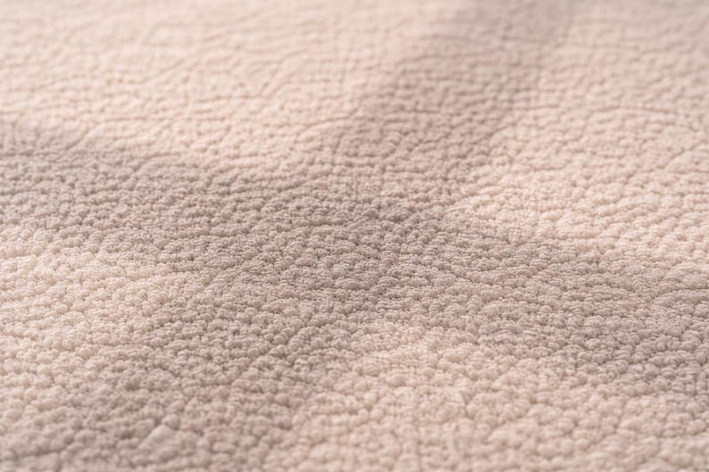 K&ouml;nnte beinhalten: Nahaufnahme eines strukturierten, hellbeigen Stoffes. Die Oberfl&auml;che hat ein weiches, pl&uuml;schartiges Aussehen mit einem Muster aus kleinen, erhabenen Unebenheiten. Die Beleuchtung erzeugt subtile Schatten, die die Textur hervorheben.