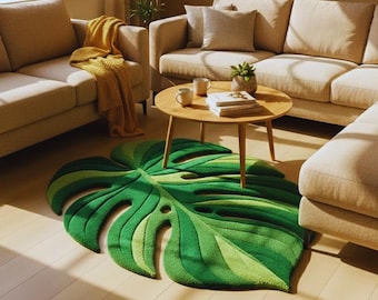 Op de afbeelding: Een groene monstera blad-vormige vloerkleed in een woonkamer. Het kleed heeft verschillende tinten groen, wat een textuur creëert. De kamer bevat beige banken, een houten salontafel en potplanten, met natuurlijk licht.