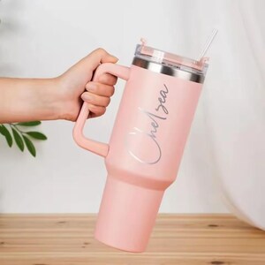 May include: A light pink insulated tumbler with a handle and a straw. The tumbler has a silver lid and the name "Chelsea" in a cursive font. The tumbler is held by a hand and sits on a wooden surface.