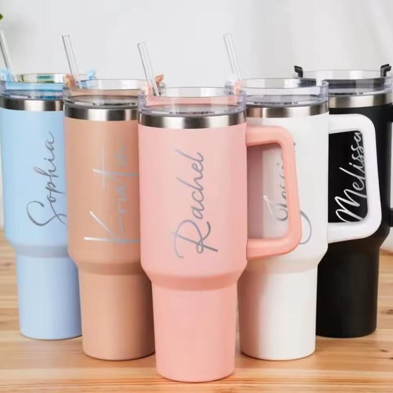 Op de afbeelding: Een verzameling gepersonaliseerde bekers in pasteltinten, waaronder lichtblauw, perzik en roze. Elke beker heeft een zilveren naamopschrift en een doorzichtige rietje. De bekers hebben een handvat en een zilveren rand.