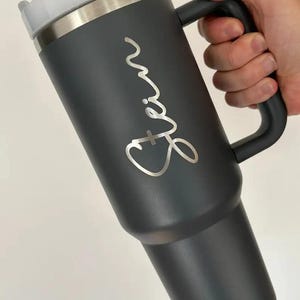 Op de afbeelding: Een donkergrijze ge&iuml;soleerde beker met een handvat, een zilveren rand en een wit deksel. De naam "Erin" staat in zilveren letters op de zijkant. Ontworpen om dranken warm of koud te houden.