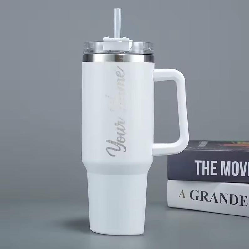 Op de afbeelding: Witte ge&iuml;soleerde beker met een handvat, een zilveren rand en een doorzichtig rietje. De tekst "Your name" staat verticaal op de zijkant. De beker staat naast twee boeken met de titels "THE MOVIE" en "A GRANDE".