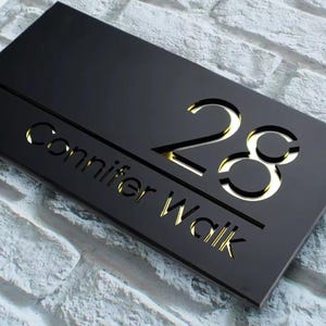 以下が含まれることがあります： 黒い長方形の表札。 金色の数字「28」と「Connifer Walk」の文字が刻まれています。 明るい灰色のレンガの壁に取り付けられています。