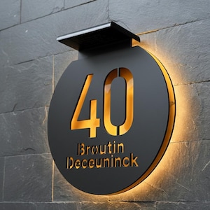 May include: A black, illuminated address sign with the number "40" in a cutout design, and the names "Broutin Deceuninck" below. The sign is mounted on a dark gray stone wall and has a built-in light.