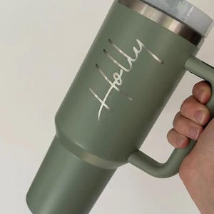Op de afbeelding: Een saliegroene ge&iuml;soleerde beker met een handvat en een doorzichtig deksel. De naam "Holly" is in zilveren letters gegraveerd. De beker is ontworpen om dranken warm of koud te houden.