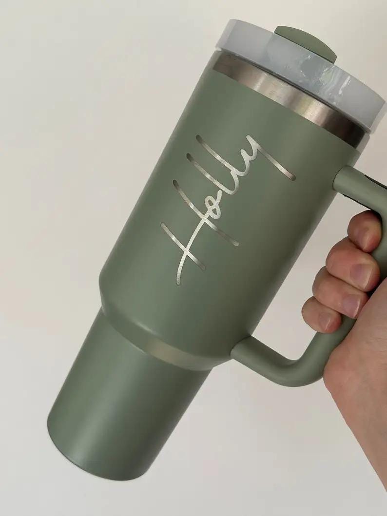 Op de afbeelding: Een saliegroene ge&iuml;soleerde beker met een handvat en een doorzichtig deksel. De naam "Holly" is in zilveren letters gegraveerd. De beker is ontworpen om dranken warm of koud te houden.
