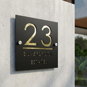 May include: Black square address sign with gold-colored numbers "23" and the text "SILVERBROOK ESTATES". The sign is mounted on a light gray concrete wall with two silver-colored screws.