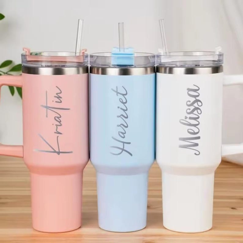 Op de afbeelding: Drie gepersonaliseerde bekers in pastelkleuren: roze, lichtblauw en wit. Elke beker heeft een zilveren deksel, handvat en rietje. De namen "Kristin", "Harriet" en "Melissa" staan verticaal op elke beker.