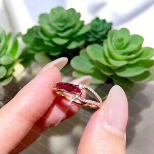 以下が含まれることがあります： ローズゴールドのリング。長方形の深い赤色のルビーがチャンネルセッティングで横向きに配置され、その隣には小さな透明なパヴェダイヤモンドが埋め込まれたバイパススタイルのバンドセグメントがあり、リングの中心に向かって細くなっています。