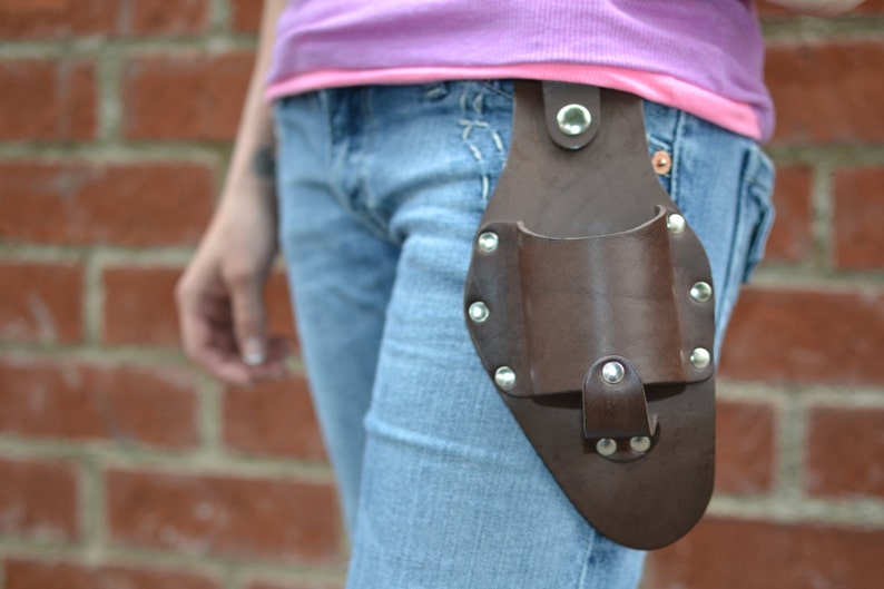 Op de afbeelding: Een bruine leren gereedschapsriem met een metalen gesp en klinknagels. De riem wordt gedragen door iemand die een blauwe jeans draagt.
