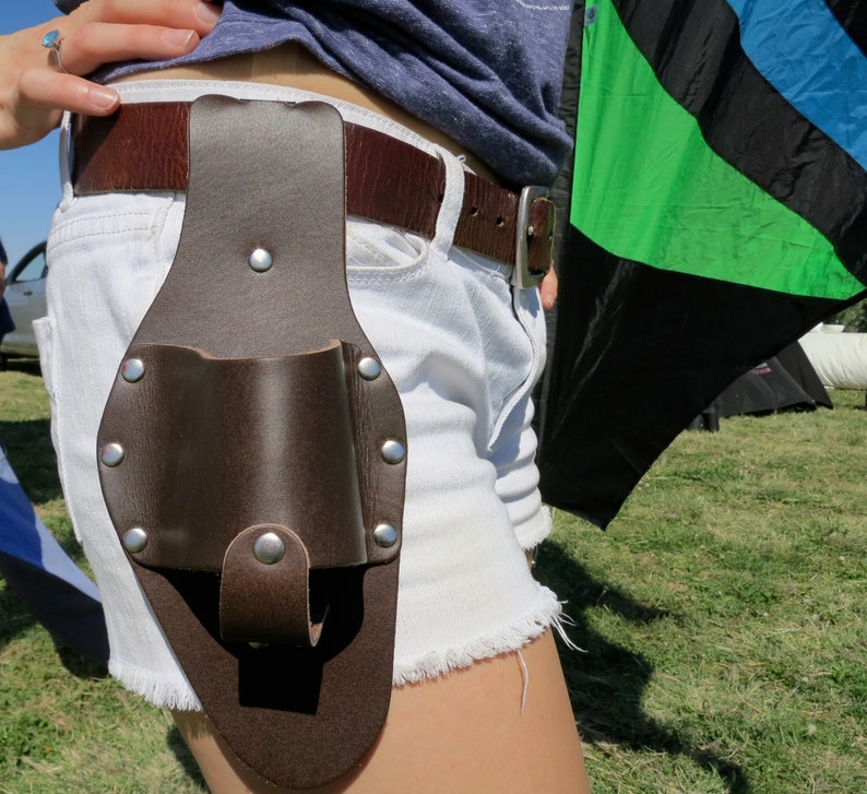 Op de afbeelding: Een bruine leren gereedschapshouder met een metalen gesp en klinknagels. De houder is bevestigd aan een riem en wordt gedragen op de heup.