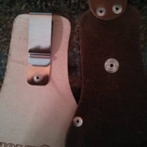 Op de afbeelding: Twee leren holster voor een zakmes of klein gereedschap. Een holster is van bruin leer met een metalen drukknoop sluiting. De andere holster is van lichtbruin leer met een metalen clip. Beide holster hebben het woord "HOLSTER" in het leer gegraveerd.