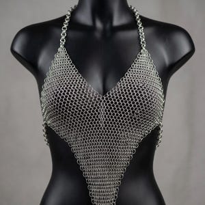 Op de afbeelding: Een zilveren maliënkolder haltertop afgebeeld op een zwarte mannequin. De top is gemaakt van kleine, in elkaar grijpende metalen ringen, die een driehoekige vorm vormen die de borst en een halternek bedekt. Het metaal heeft een glanzende, reflecterende kwaliteit.