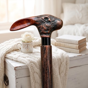 May include: A walking stick featuring a handle shaped like a rabbit head with a long, polished brown ear and a textured, dark bronze-toned finish. The shaft of the stick has a matching dark, weathered wood-grain appearance with a black collar.