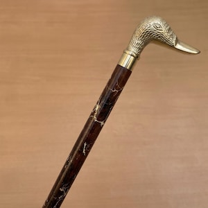 Pode incluir: Bengala decorativa com uma pega em forma de cabeça de pato em latão. A pega apresenta detalhes de penas texturizadas e um bico longo. O veio é castanho escuro com veios dourados. Uma faixa de latão une a pega ao veio.