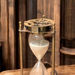 May include: An antique hourglass with a brass frame and a detailed top piece. The glass bulb contains light-coloured sand, and the hourglass is set against a wooden background. A classic timekeeping device.