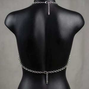 May include: A silver-colored body chain necklace. The necklace is composed of small, linked metal rings that form a chain. The chain drapes across the back of a black mannequin, with a clasp closure at the neck and waist. The chain is designed to be worn as a fashion accessory.