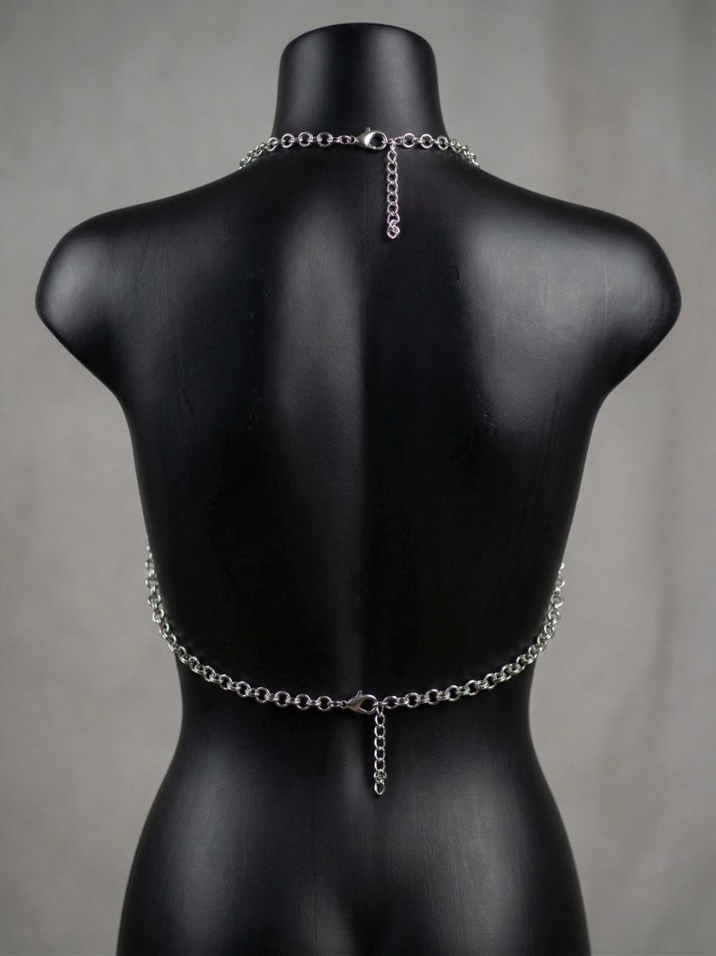 May include: A silver-colored body chain necklace. The necklace is composed of small, linked metal rings that form a chain. The chain drapes across the back of a black mannequin, with a clasp closure at the neck and waist. The chain is designed to be worn as a fashion accessory.